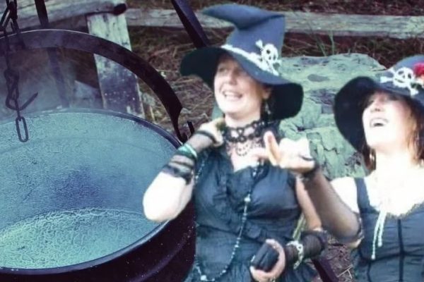 2 women dressed as witches standing near a cauldron.